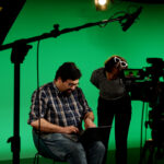 Two people stand in front of a green screen, ready for digital editing and insertion of a background image.