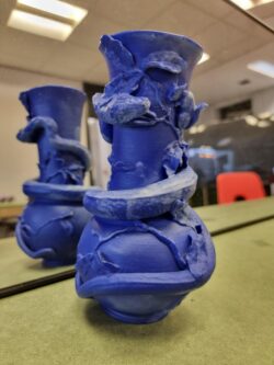 A 3d printed blue vase. The vase features sculpted plants and a snake wrapped around the vase.